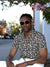 A man in sunglasses and an ID Menswear Floret Short Sleeve Printed Rayon Shirt sits on a sidewalk by a white wall and pink flowers, with trees and buildings in the background.