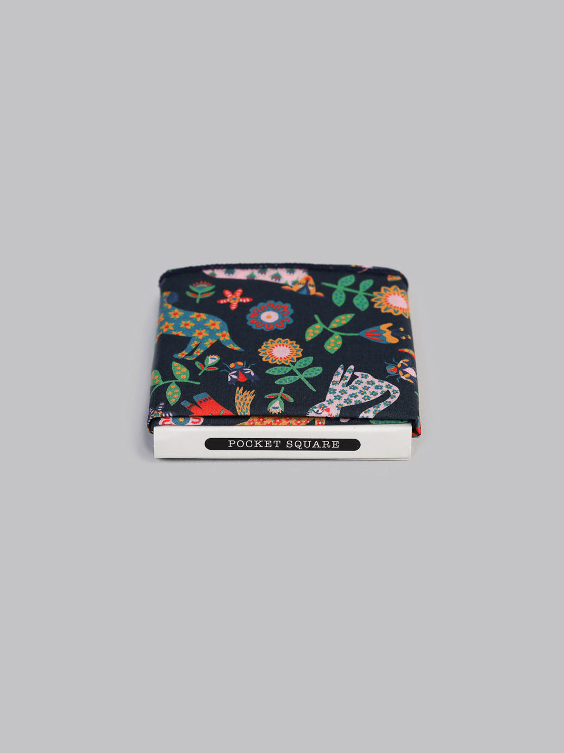 A folded Liberty™ Folk Tale Pocket Square by ID Menswear, featuring a colorful floral print on dark Tana Lawn cotton, is displayed in white packaging labeled POCKET SQUARE against a plain gray background.