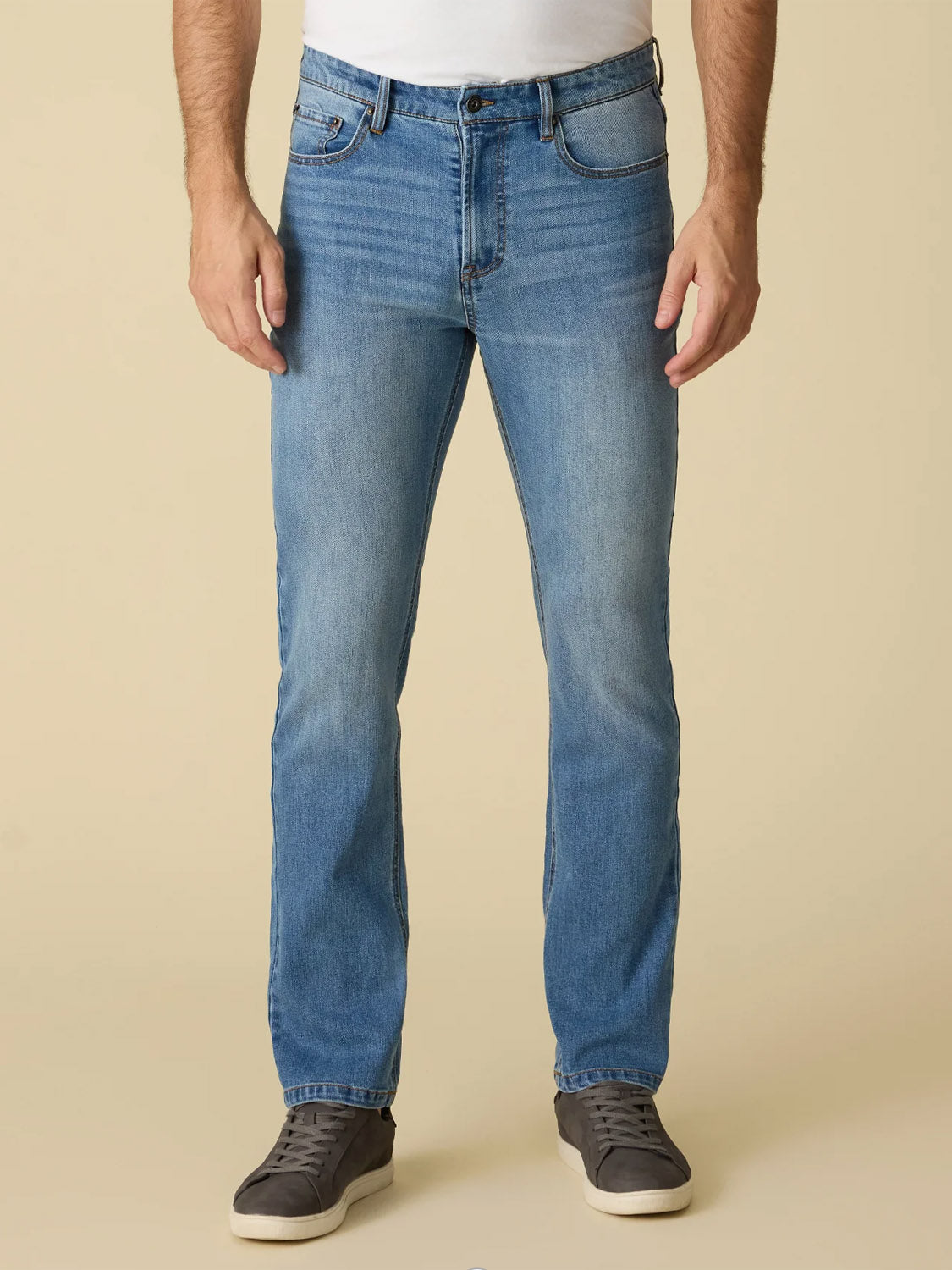 A person wearing Flag and Anthem’s Fontana Cooling Jean-Nashville Straight, teamed with a white shirt and gray sneakers, stands against a plain beige background.