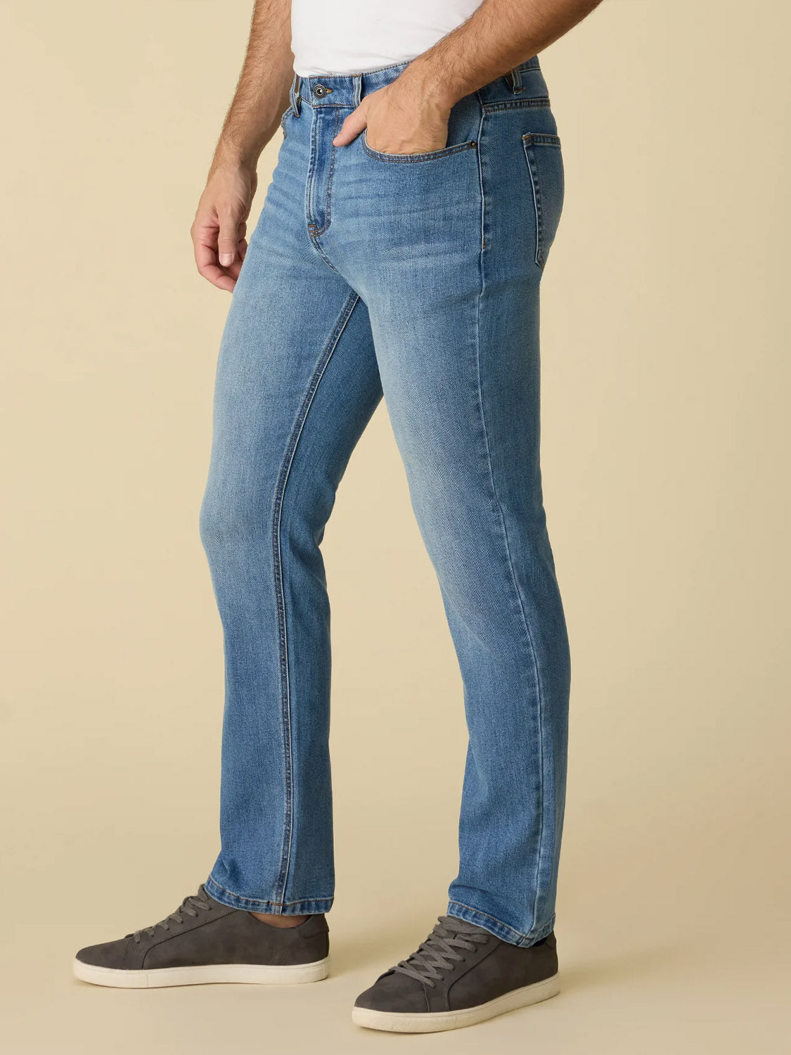 Wearing Flag and Anthem’s Fontana Cooling Jean-Nashville Straight in light blue, paired with a white shirt and gray sneakers, a person poses with a hand in their pocket against a beige background, lower body only visible.