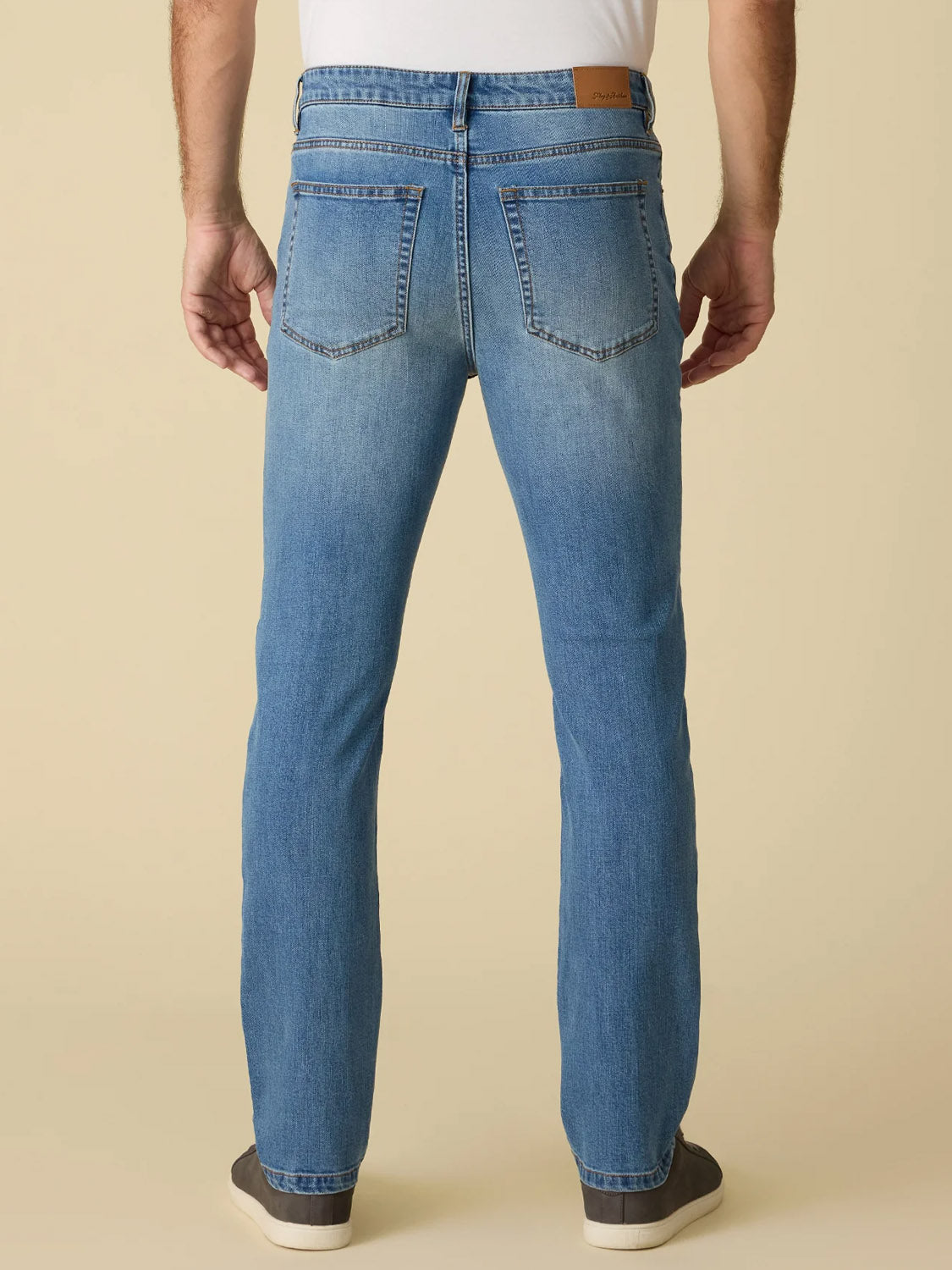 A person wearing Flag and Anthem’s Fontana Cooling Jean-Nashville Straight, seen from the back with hands at their sides. Light blue, breathable straight-leg jeans have two back pockets, a brown waistband patch, black shoes, and a white shirt.