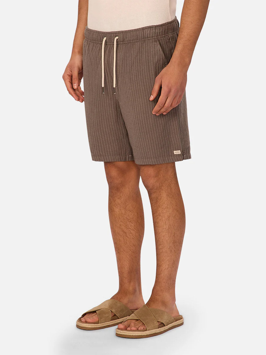 A person wearing The Fontera Short by Industrie, featuring brown pinstripes and a drawstring waist, stands against a plain white background. Only the lower body and tan suede sandals are visible.