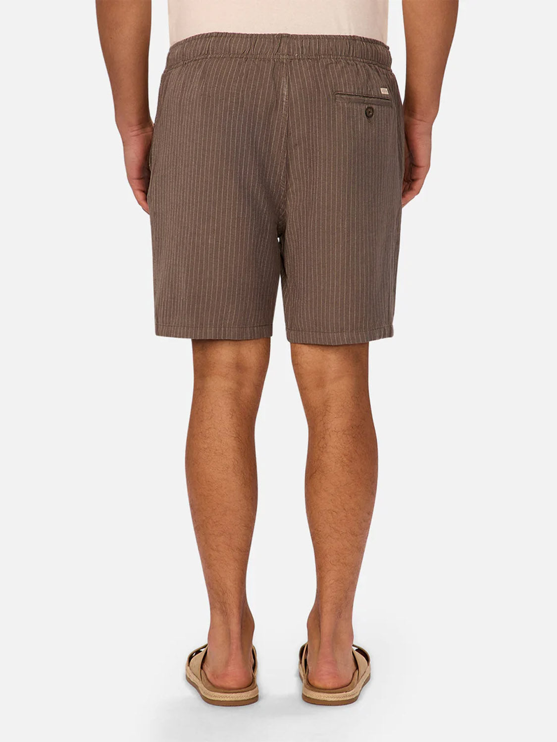 A person is shown from the back wearing Industrie’s The Fontera Short in brown pinstripe, paired with a light shirt and flip-flops, standing against a white background. These breathable shorts offer comfort and style for warm weather.