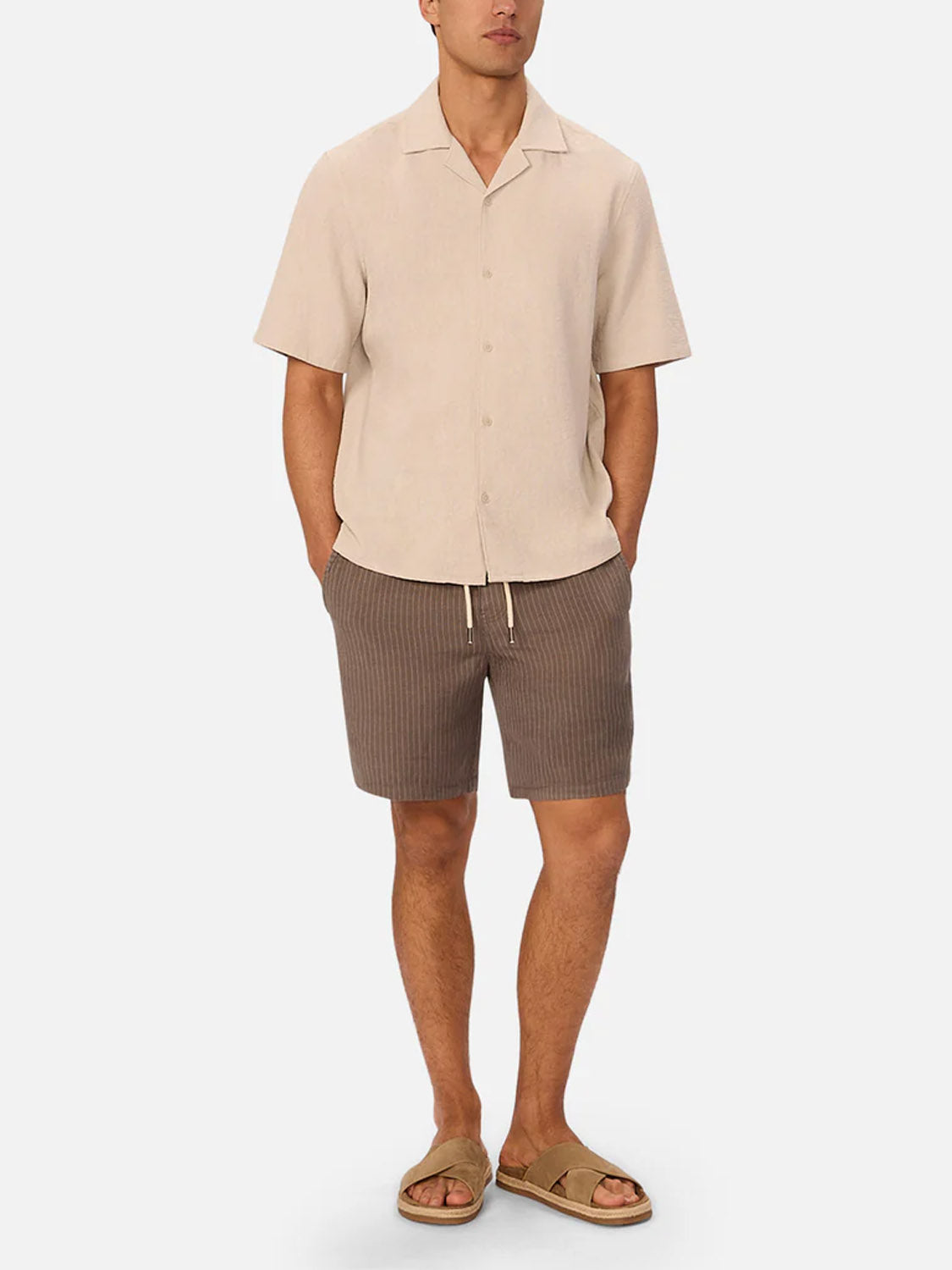 A man stands against a plain white background wearing The Fontera Short by Industrie, styled with a short-sleeve beige button-up shirt and tan slip-on sandals.