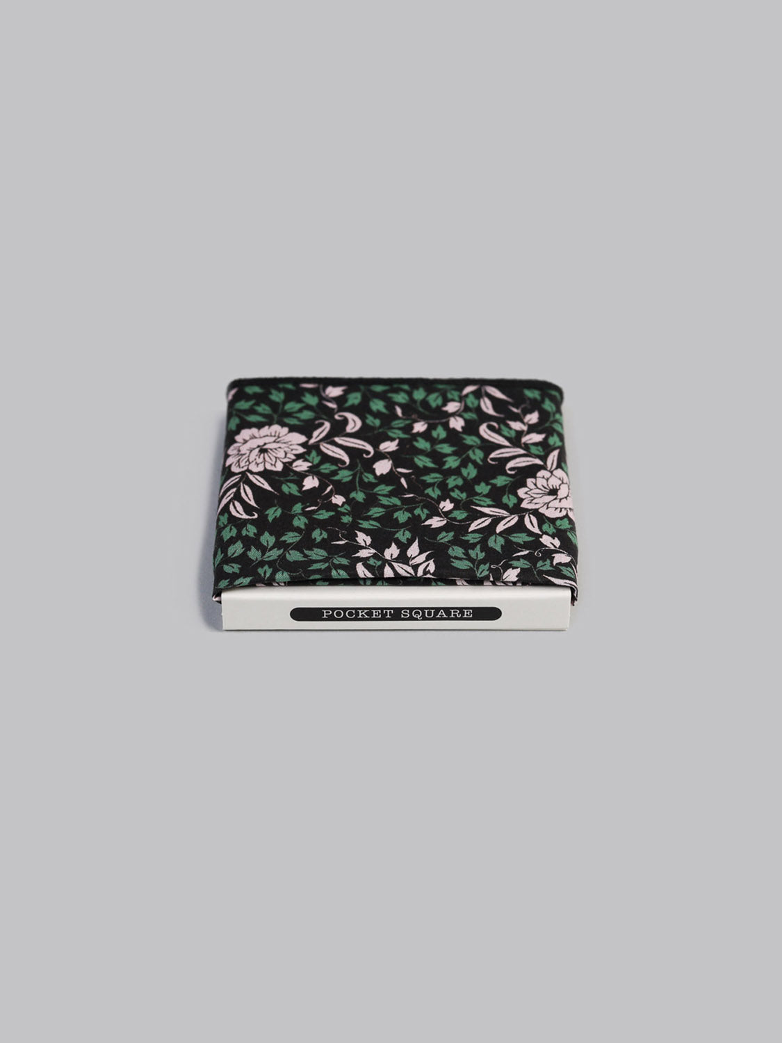 A folded Liberty™ Franklin Pocket Square by ID Menswear, handmade in Brooklyn from Tana Lawn cotton, features a black background with white and pink flowers and green leaves. It comes boxed with POCKET SQUARE on the label atop a light gray surface.