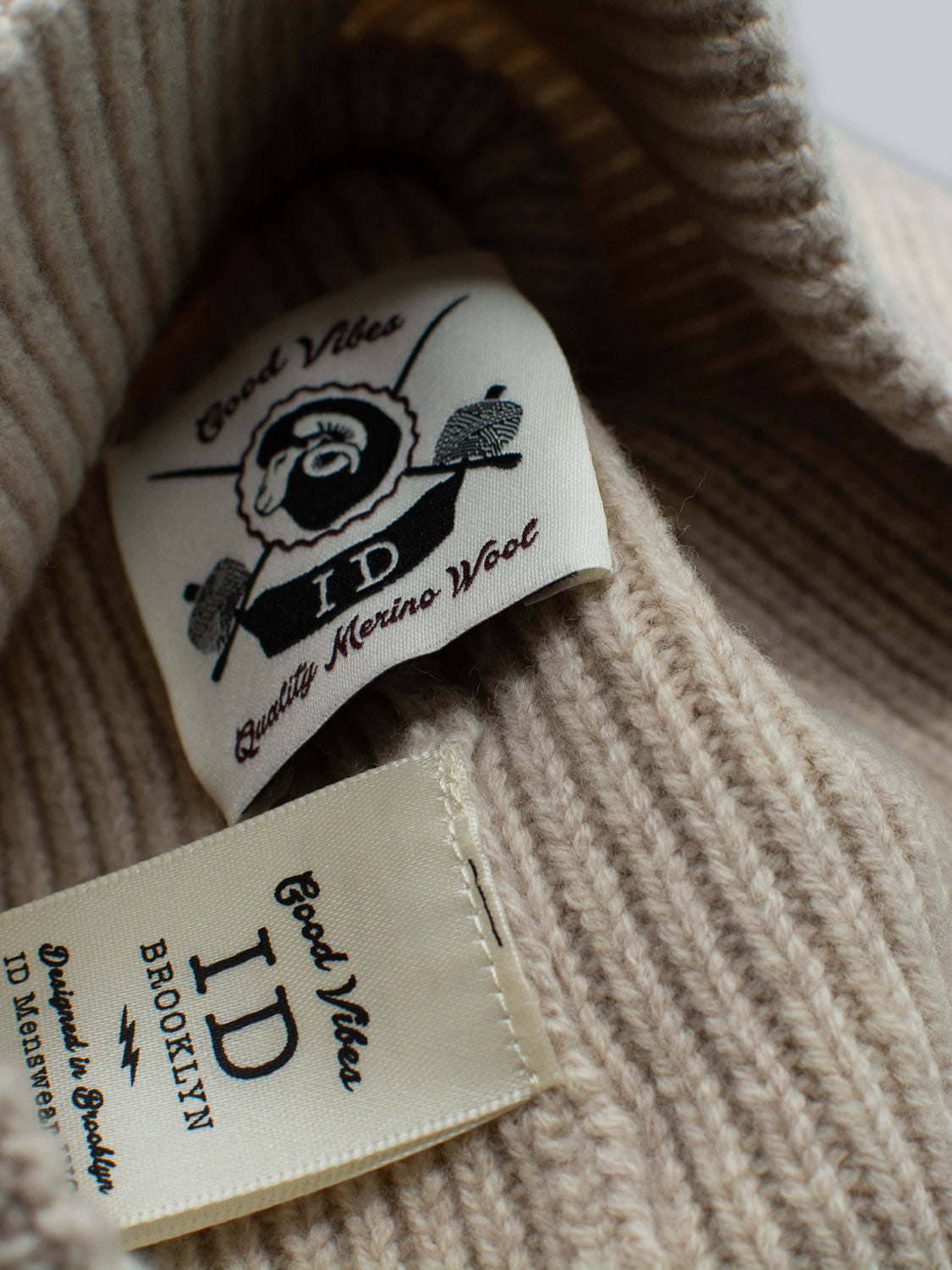 Close-up of beige ribbed wool fabric featuring two labels. One reads ID Menswear 100% Merino Wool with a bearded man graphic; the other says ID Menswear Brooklyn with care instructions, making the Fuji Merino Wool Beanie an ideal winter companion.