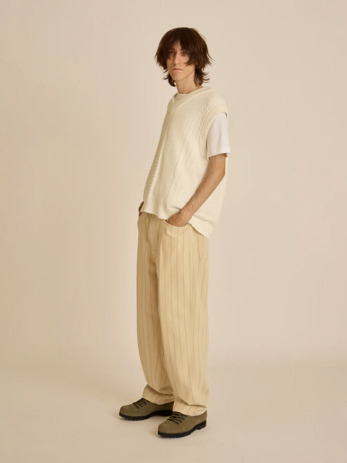 Against a plain backdrop, a person sports an Obey Garret Sweater Vest over a white tee, paired with beige wide-legged pants and gray shoes. Crafted from 100% cotton, this outfit combines comfort and style. Hands are tucked into pockets as they display medium-length hair.