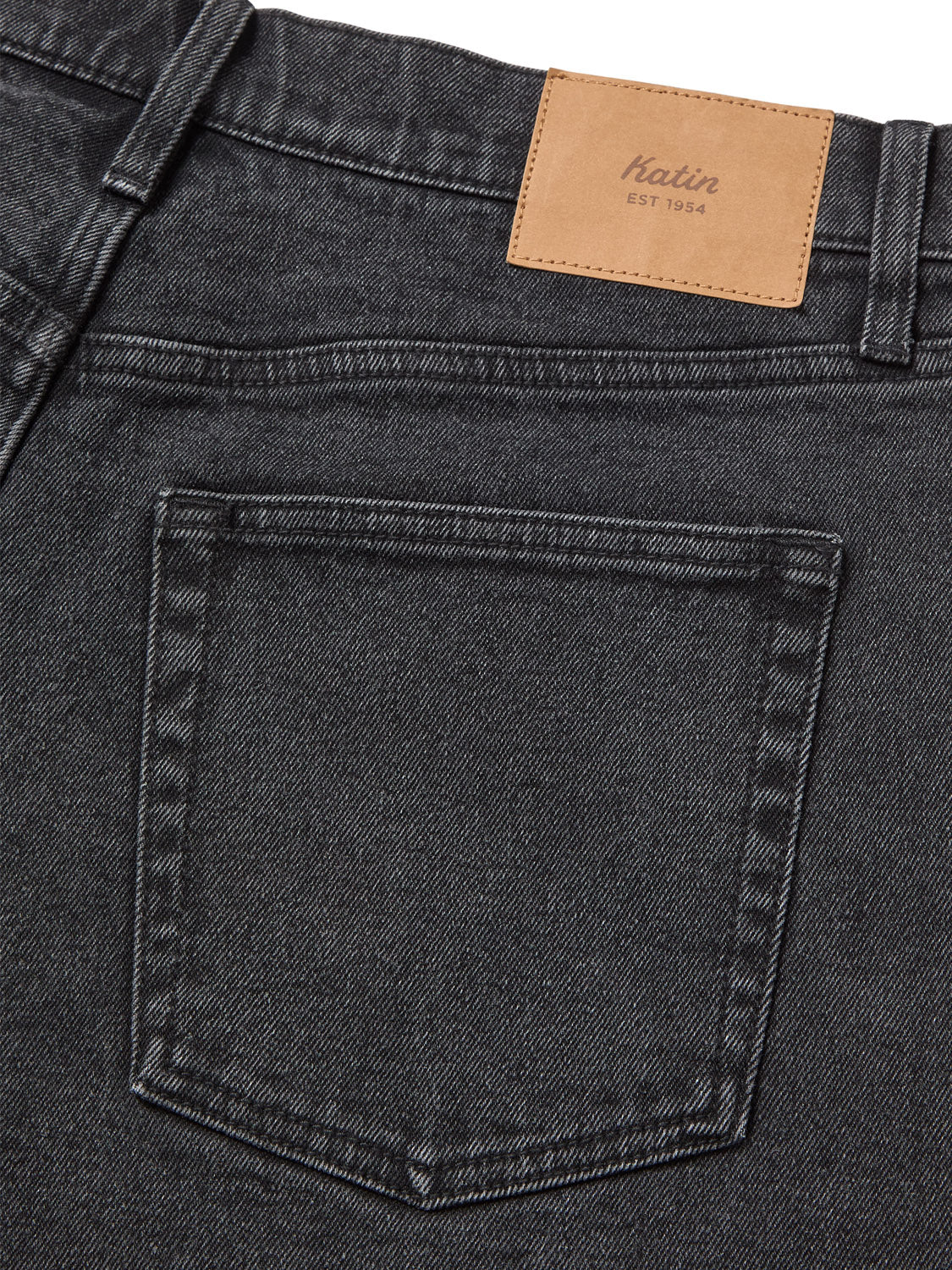 A close-up of the back pocket of relaxed fit dark gray jeans highlights a brown leather patch above, branded Katin EST 1954. The classic design with visible stitching is reminiscent of the Katin George 64 Short - 20 collection.