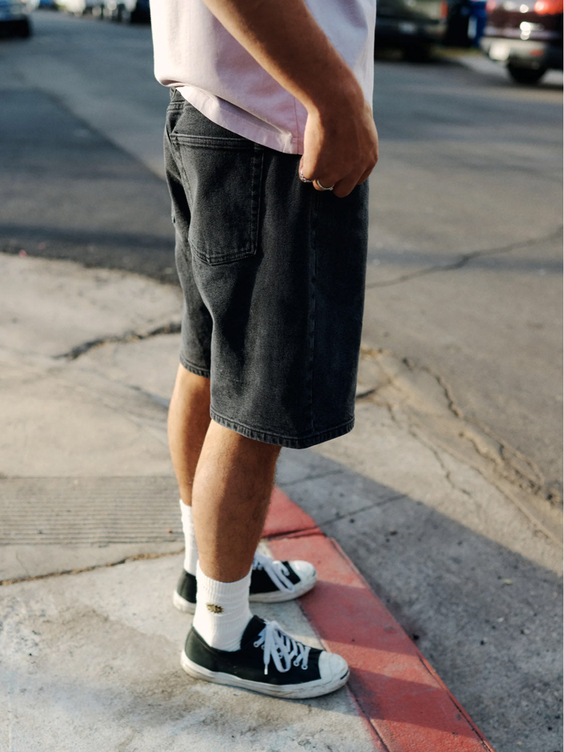 A person in a light pink shirt and Katin George 64 Short - 20 in classic wash stands on a street corner. The relaxed fit, complemented by white socks and black sneakers, is illuminated by natural light with parked cars visible against the concrete background.
