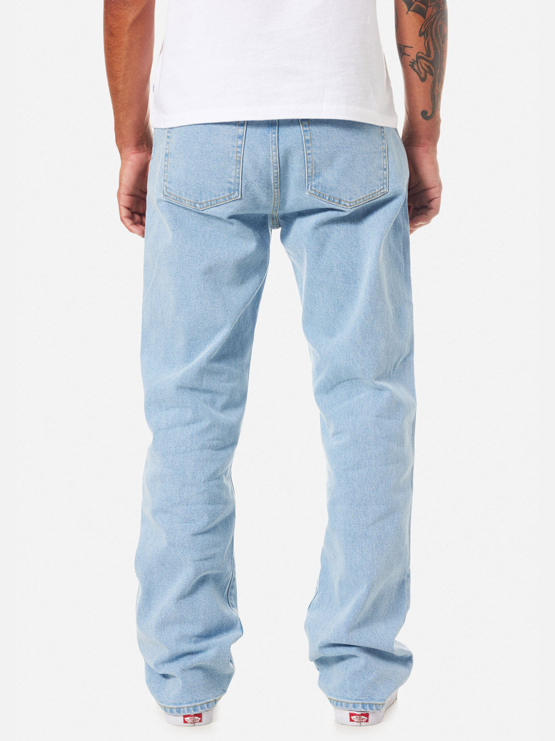 A person wearing Katin George 64 Relaxed Pants in light blue, a white t-shirt, and white sneakers stands with their back to the camera. A tattoo is visible on their right arm.
