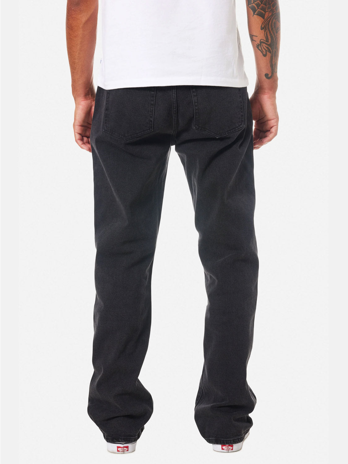 A person with a tattooed arm faces away from the camera, wearing a white T-shirt, white sneakers, and Katin George 64 Relaxed Pants with 5-pocket styling and back pockets. The relaxed-fit jeans are set against a plain white background.