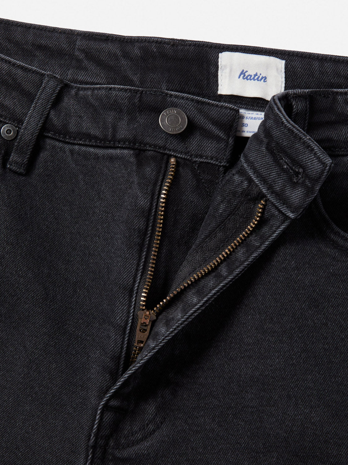 Close-up of Katin George 64 Relaxed Pants in black denim, featuring a partially open brass zipper, button closure, and 5-pocket styling. The jeans are laid flat on a white background with the inside label showing the Katin brand.