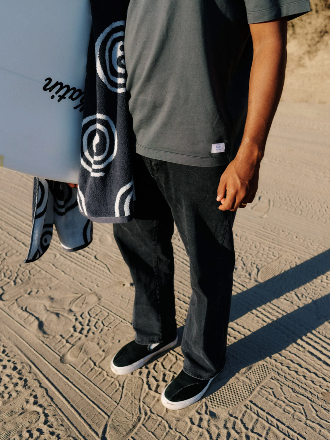 A person stands on sand, holding a surfboard and a patterned towel. They wear Katin’s George 64 Relaxed Pants, a dark t-shirt, and black slip-on shoes. Only their lower half is shown.