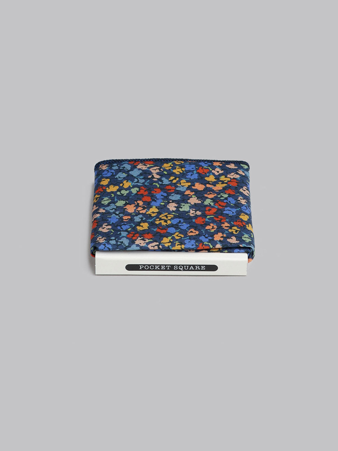 A folded Liberty™ Gizmo Pocket Square by ID Menswear, featuring a colorful floral design on dark Tana Lawn cotton, rests on a white box labeled POCKET SQUARE against a gray background. Handmade in Brooklyn.