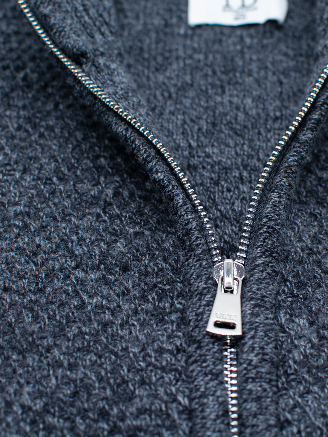 Close-up of the Glasgow Zip Up Sweater by ID Menswear in textured dark gray merino wool, featuring a silver metal zipper partially unzipped to reveal the fine knit pattern of the lightweight material.
