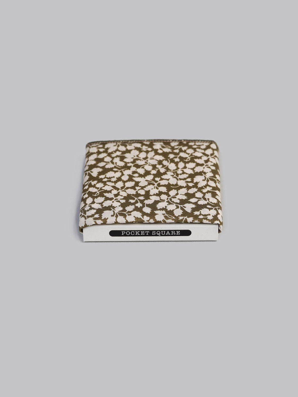 A folded Liberty™ Glen Jade Pocket Square by ID Menswear, featuring a brown and white floral pattern on Tana Lawn cotton, rests atop a white POCKET SQUARE box against a plain light gray background.