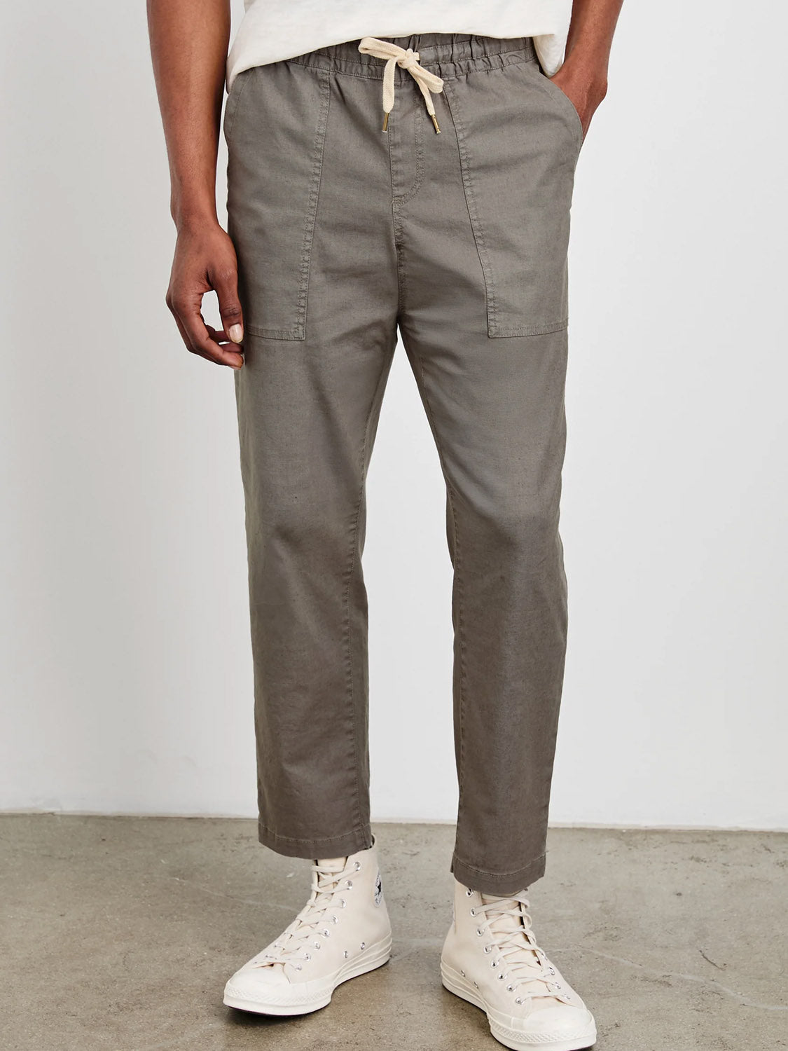 A person stands on a concrete floor in gray Rails Gobi Pants featuring sustainable buttons, front patch pockets, and a slim fit, paired with white high-top sneakers. Only the lower half of their body is visible against a plain background.