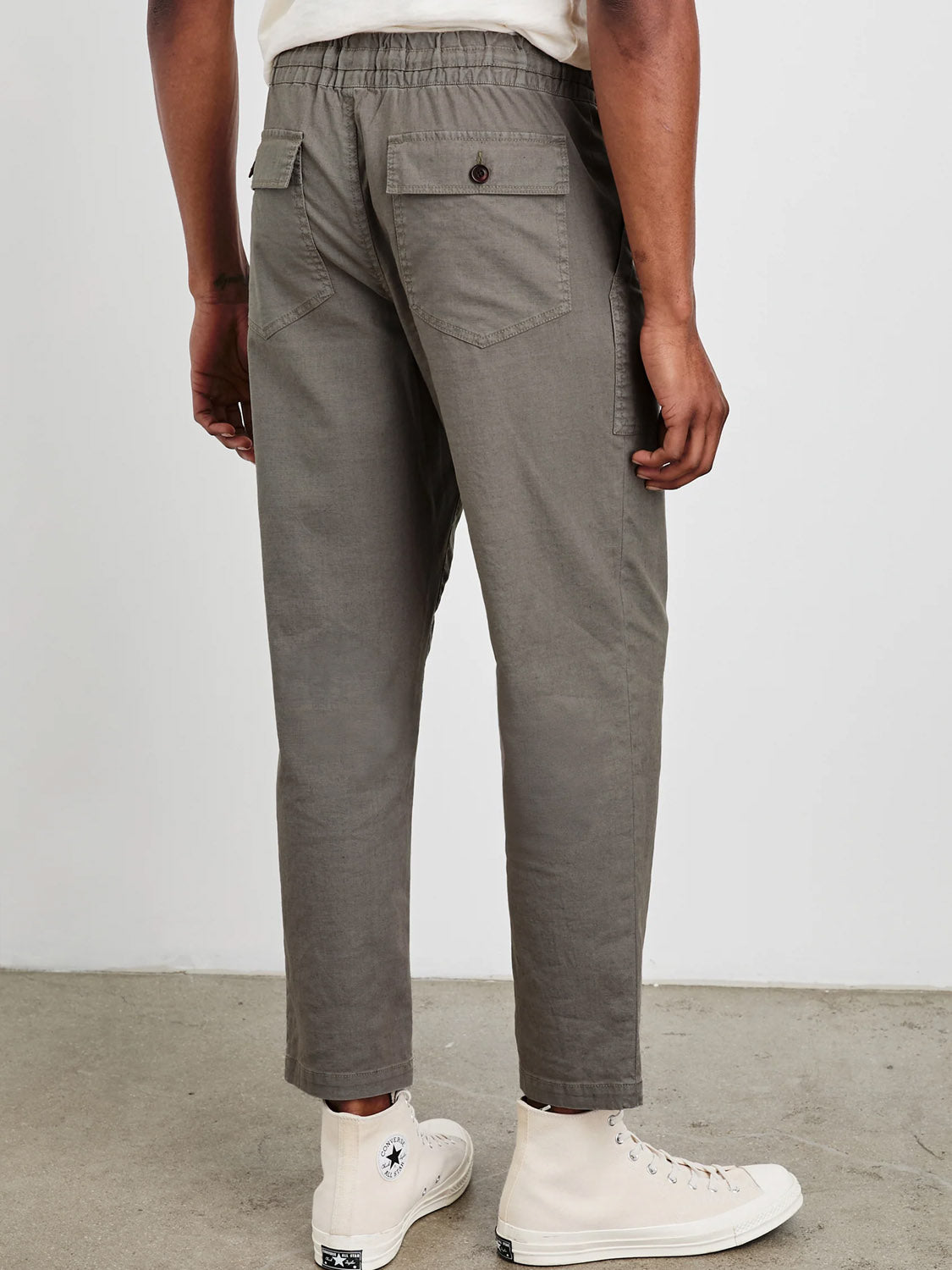 The image shows a person facing away, wearing Rails Gobi Pant in olive, featuring a slim fit with sustainable buttoned back pockets, paired with white high-top sneakers on a concrete floor.