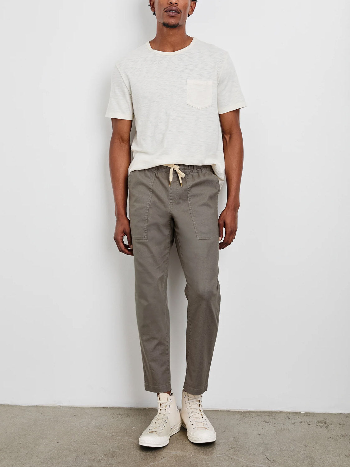 A person stands against a white wall, wearing a light gray T-shirt and Rails Gobi Pant in olive-green. The pants feature linen fabric and sustainable buttons. Beige high-top sneakers complement the slim-fit appearance, perfectly balancing casual and relaxed vibes.
