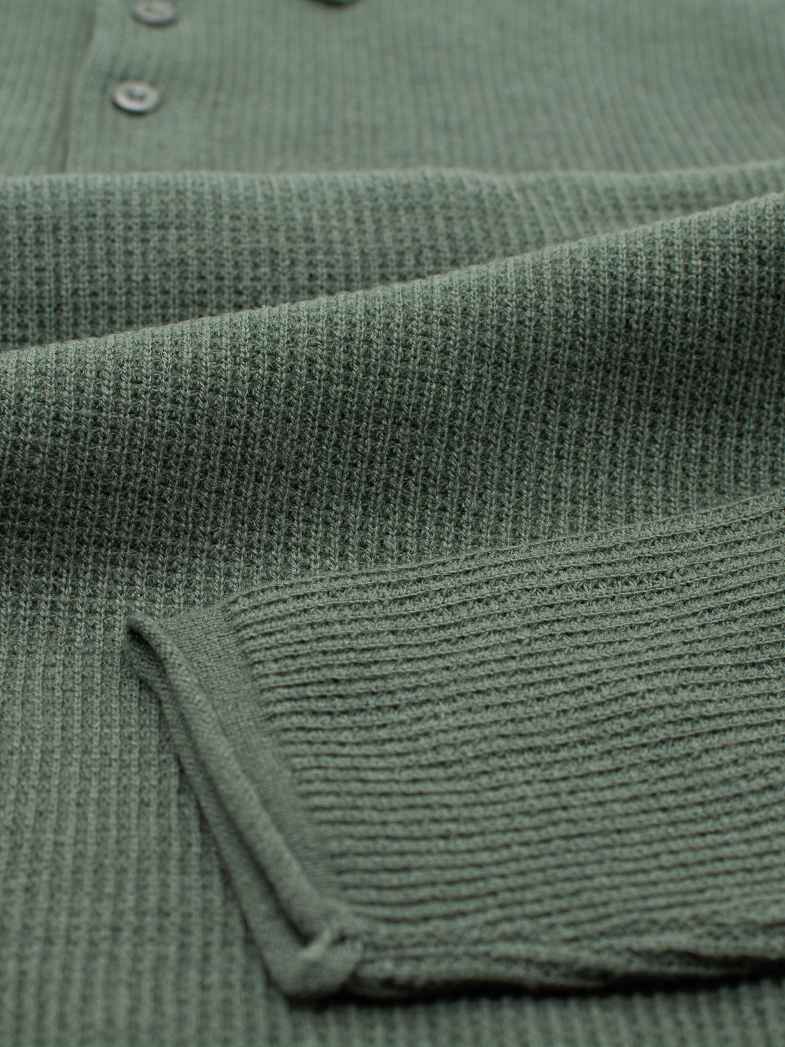 Close-up of a green waffle-knit fabric from the Gothenberg Merino Sweater Polo by ID Menswear, showing buttons and a folded piece atop. The texture showcases the knit pattern and softness, embodying casual elegance in every stitch.