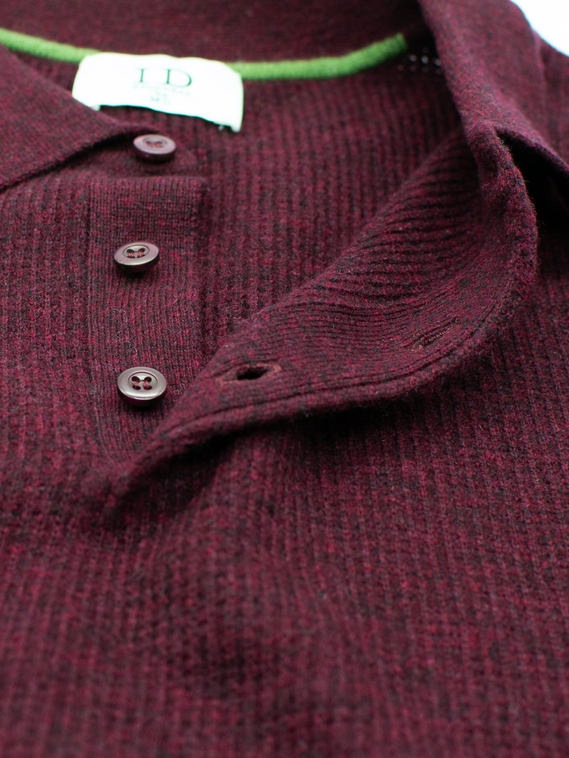 Close-up of the Gothenberg Merino Sweater Polo by ID Menswear. This long sleeve burgundy knit shirt features a textured pattern, collar, brown buttons down the front, and green trim inside. A partially visible label with brand logo is sewn on the inside, exuding casual elegance.