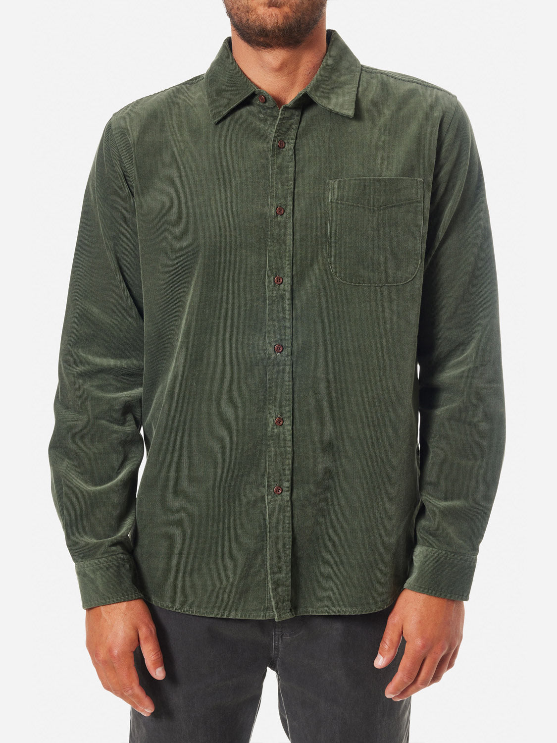 A person stands against a plain white background wearing a dark green Katin Granada Shirt from the fall collection, featuring brown buttons and a chest pocket. Crafted from soft cotton corduroy, the shirt pairs seamlessly with their dark pants.