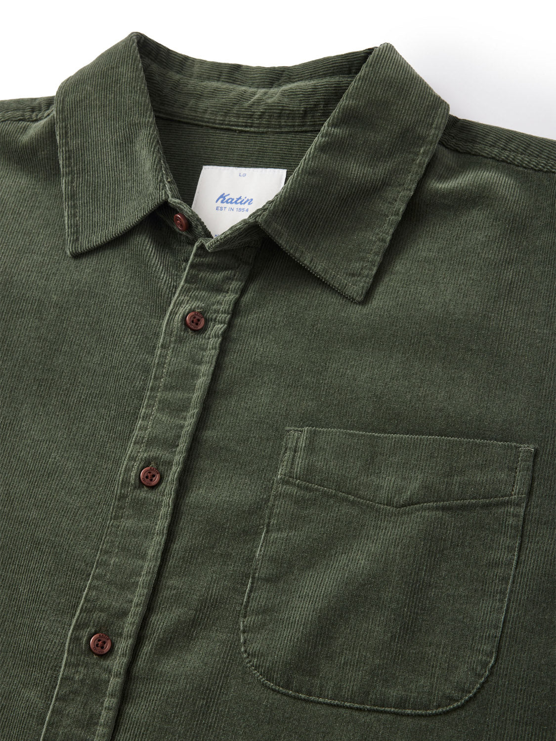 Close-up of the Katin Granada Shirt from the fall collection, made using green cotton corduroy with a collar, front pocket, and brown buttons. A white label featuring blue text is visible inside the collar. Its neatly displayed on a light background.