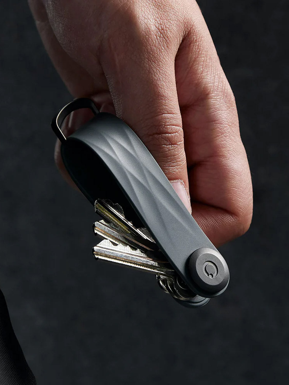 A hand holds the Orbitkeys Key Organizer Rubber Active, a sleek black key holder with several keys folded inside, multitool-style. Its durable design stands out against the dark, blurred background.