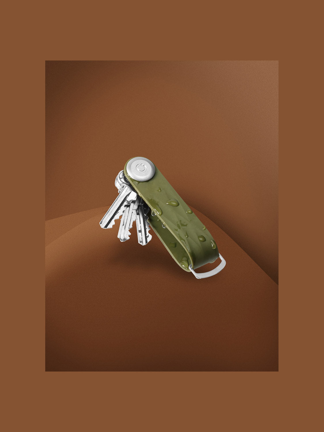 The Orbitkeys Key Organizer Rubber Active, in olive green, is displayed on a smooth brown surface with several silver keys stacked neatly inside and water droplets highlighting its durable design.
