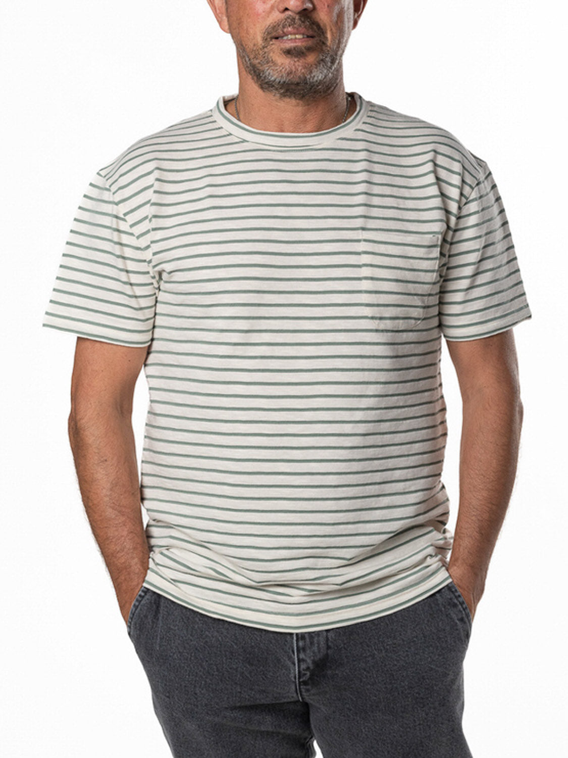 Man in La Paz Guerreiro Tee, a white and gray striped short-sleeve t-shirt made of lightweight cotton, paired with dark gray pants, standing with hands in pockets against a plain white background.