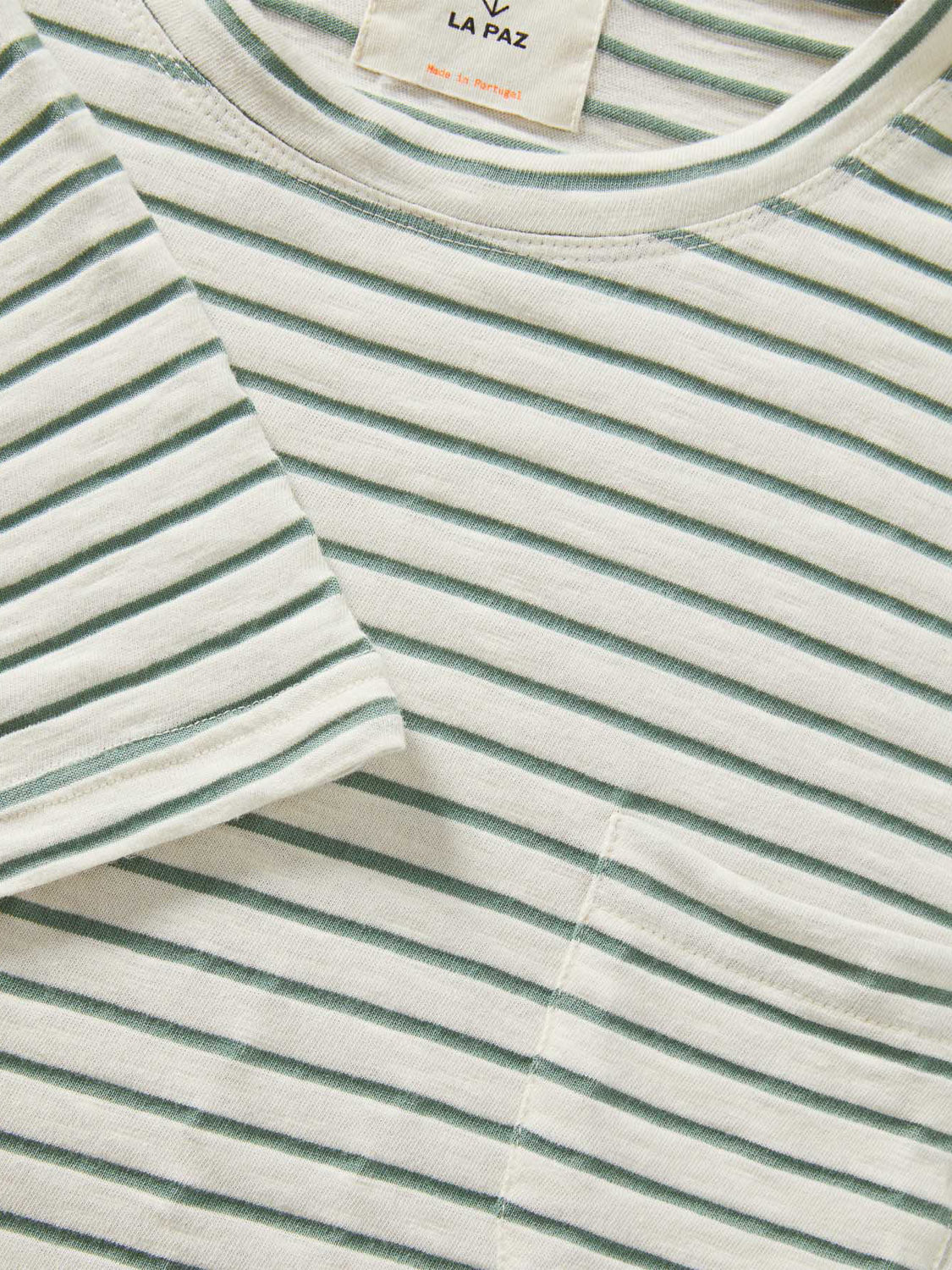 Close-up of the La Paz Guerreiro Tee, a light green and white striped T-shirt crafted from lightweight cotton. The neckline and sleeve detail are visible with the La Paz tag above. This soft tee also has a subtle side pocket, ideal for summer wear.