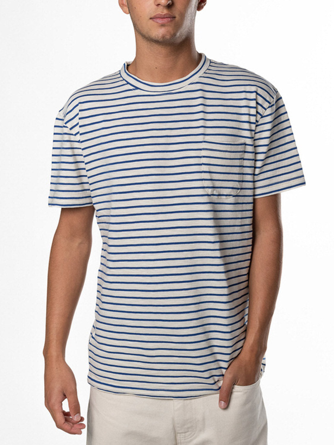 Person in a La Paz Guerreiro Tee, featuring white and blue stripes, crafted from lightweight cotton with a chest pocket, paired seamlessly with light-colored pants against a plain white background for a perfect summery look.