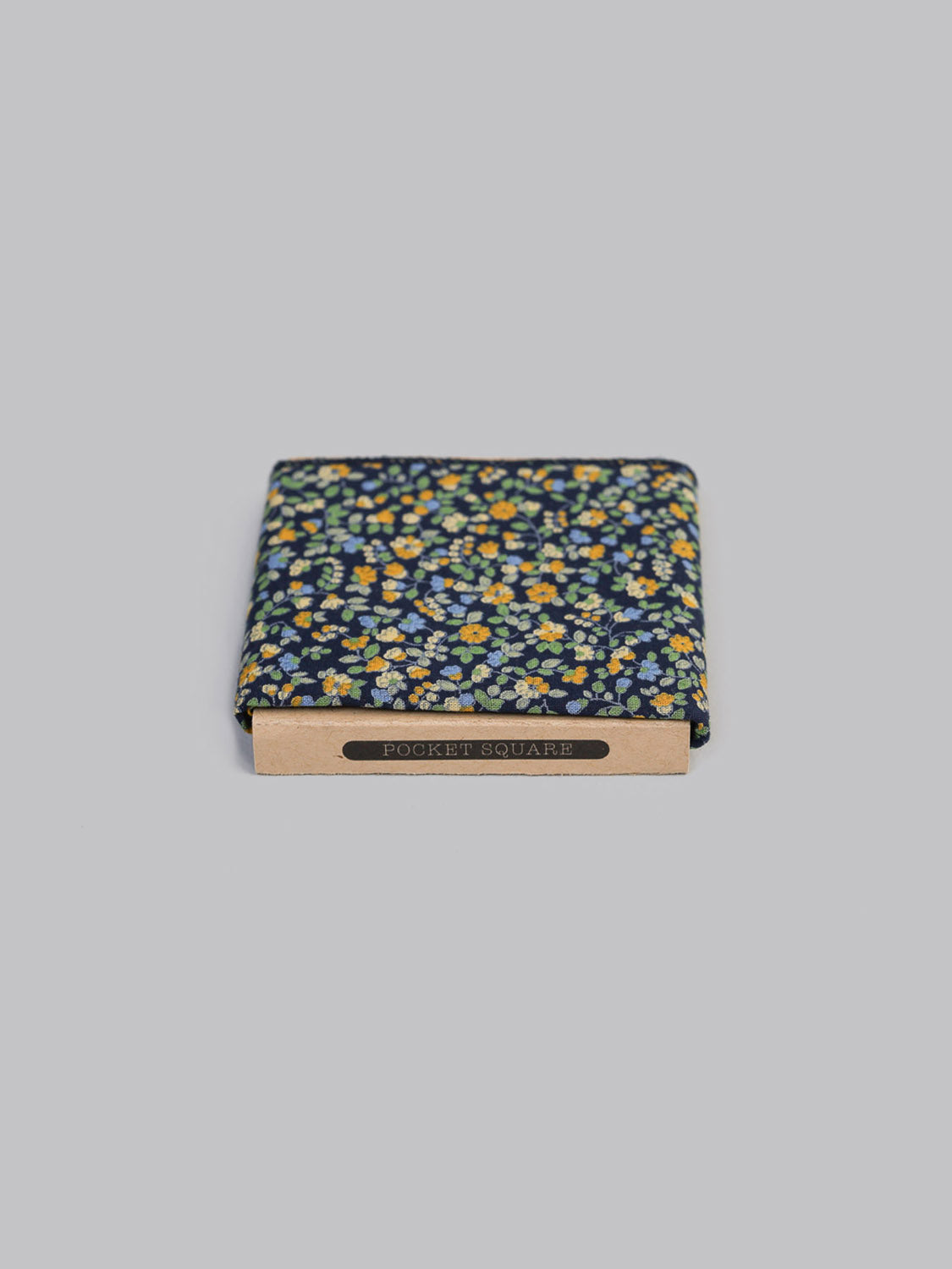 The Hana NG Pocket Square by ID Menswear is handmade in Brooklyn from 100% cotton, featuring a navy background with a yellow-green floral design. It comes folded in a cardboard sleeve labeled POCKET SQUARE on a plain light gray backdrop.