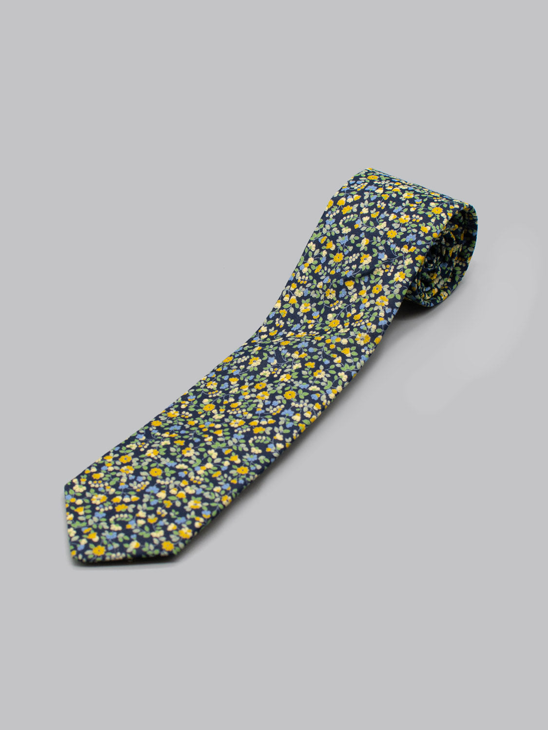 The Japanese Printed Hana NG Tie by ID Menswear features a dark background with small yellow, white, and blue floral prints. Handmade in Brooklyn from 100% cotton, its shown against a light gray surface.