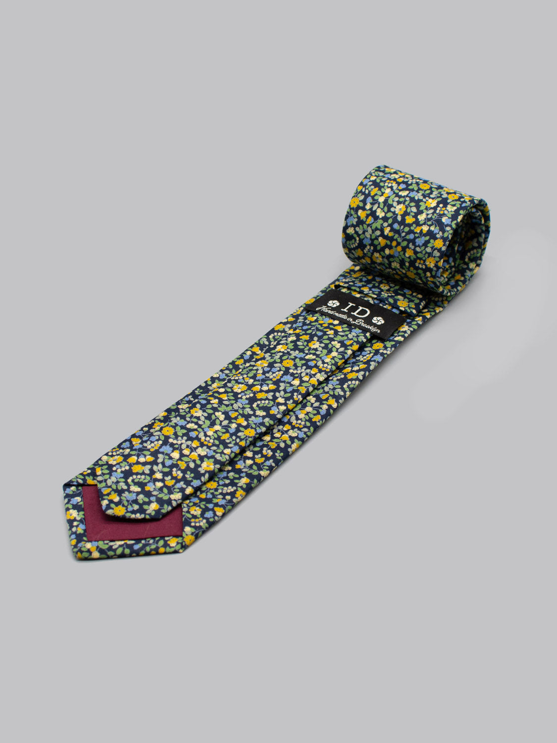 The Japanese Printed Hana NG Tie by ID Menswear is handmade in Brooklyn from 100% cotton, featuring a navy base with yellow, white, and light blue flowers, a maroon tip, and a black label near the wide end—displayed on a gray surface.