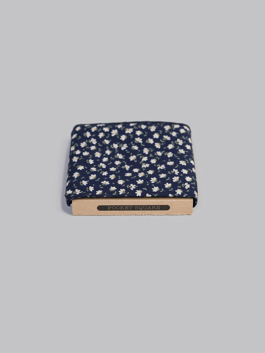 The Hana NW Pocket Square by ID Menswear features a navy Japanese floral print, handmade in Brooklyn, and comes folded in a brown holder labeled POCKET SQUARE, set against a light gray background.