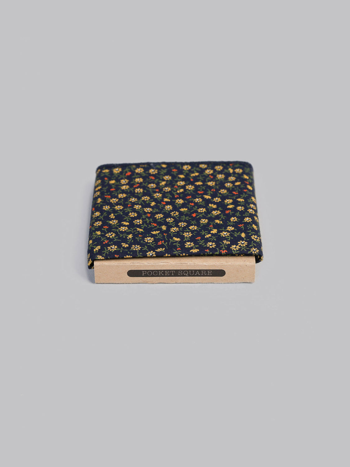 The Hana NYGR Pocket Square by ID Menswear is a handmade 100% cotton navy blue accessory with a small yellow floral pattern, shown on a light gray background and partially inside a cardboard holder labeled POCKET SQUARE.