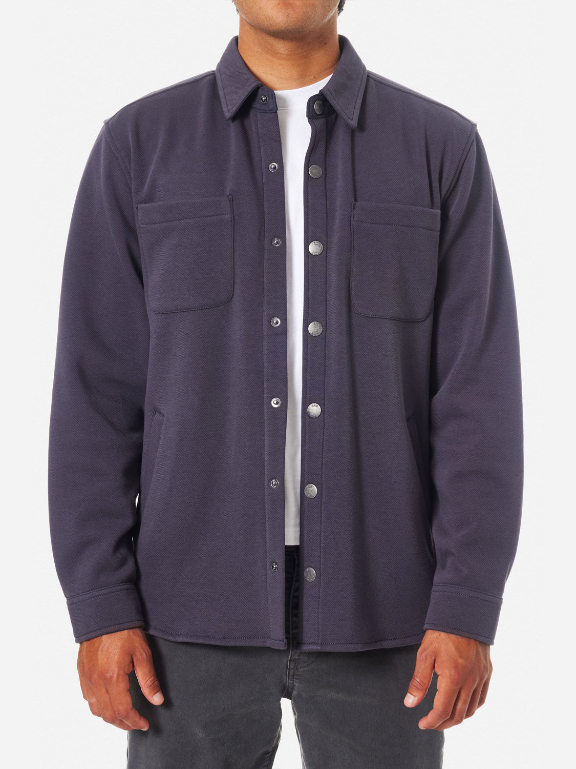 Someone is wearing a dark blue Katin Harold Fleece Jacket, a poly-cotton blend with two chest pockets and snap closures, over a white shirt. This ensemble pairs elegantly with dark pants against a plain white backdrop, highlighting the outfits classic appeal.