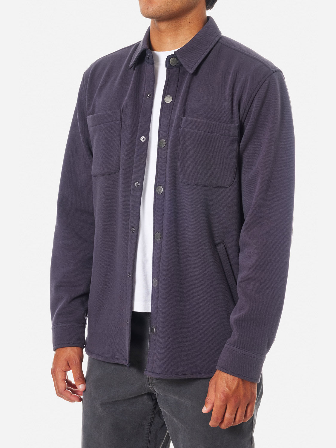 A person wearing a Katin Harold Fleece Jacket, a dark blue poly-cotton blend with snap closures and two chest pockets, stands against a white background. Their face is obscured from view in the image; beneath it, they wear a white t-shirt and dark pants.