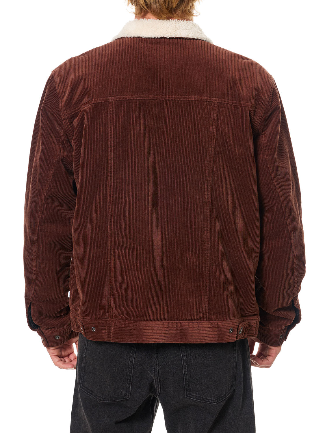 A person is seen from the back wearing the Katin Harris Corduroy Jacket, a brown cotton coat with light sherpa lining and button details at the hem, paired with dark jeans.