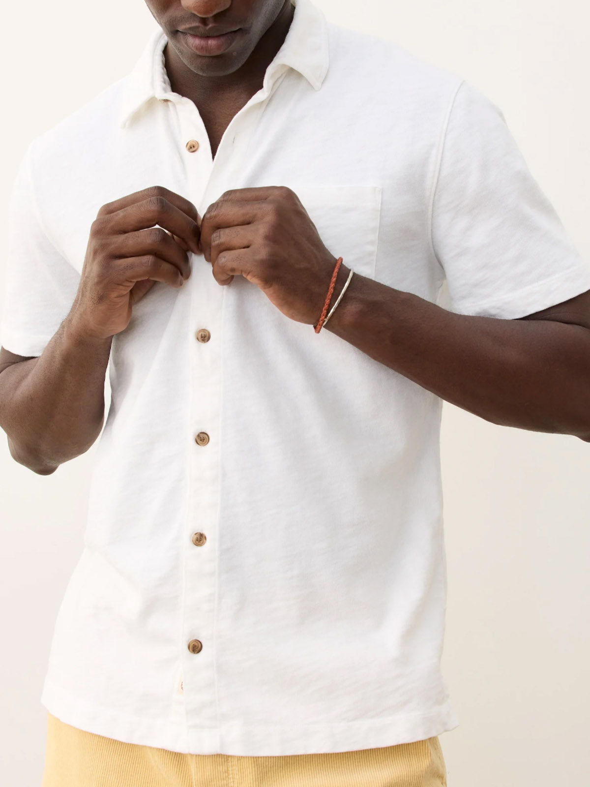 A person is buttoning up a Marine Layer Vintage Heavy Slub Button Down, a short-sleeve 100% cotton white shirt. Only their torso and hands, a thin brown bracelet, and light yellow pants are visible.