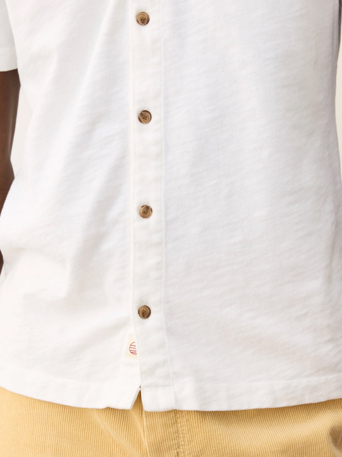 A close-up of a person wearing Marine Layer’s Vintage Heavy Slub Button Down, a garment-dyed 100% cotton white shirt with brown buttons, paired with tan corduroy pants. The shirt is partially untucked; only the torso is visible.