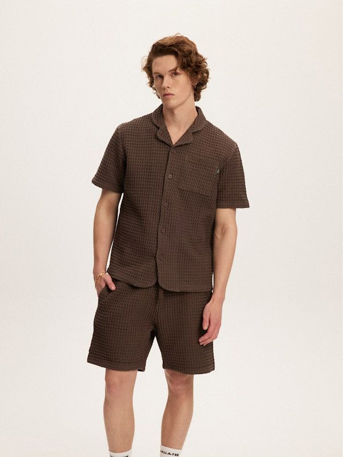 A person with wavy hair wears a matching Kuwalla Heavy Waffle Shirt and shorts set against a white background. Made from 100% cotton, the brown textured short-sleeved shirt and relaxed fit ensemble include visible white socks and partial shoes, with hands relaxed at their sides.