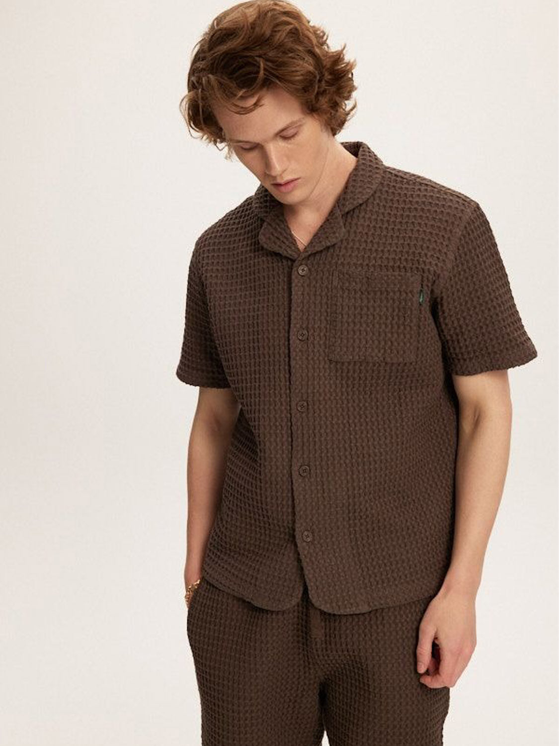 The person stands gazing down in a matching Kuwalla Heavy Waffle Shirt and pants, both dark brown with a textured design. The relaxed fit and 100% cotton fabric offer comfort. The shirt has buttons and a chest pocket, all set against a plain, light-colored background.