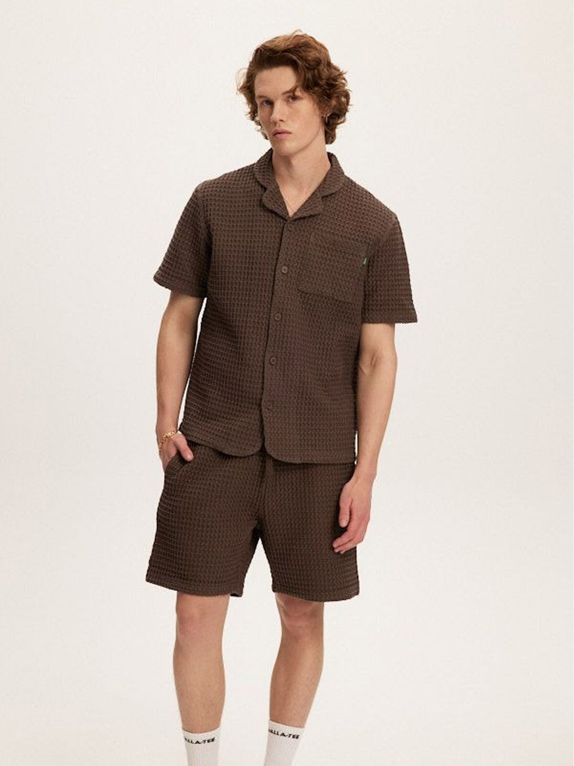 A person with wavy brown hair models the Kuwalla Heavy Waffle Short, paired with a matching brown short-sleeve button-up, standing against a white background. They have one hand in their pocket and wear a relaxed expression.