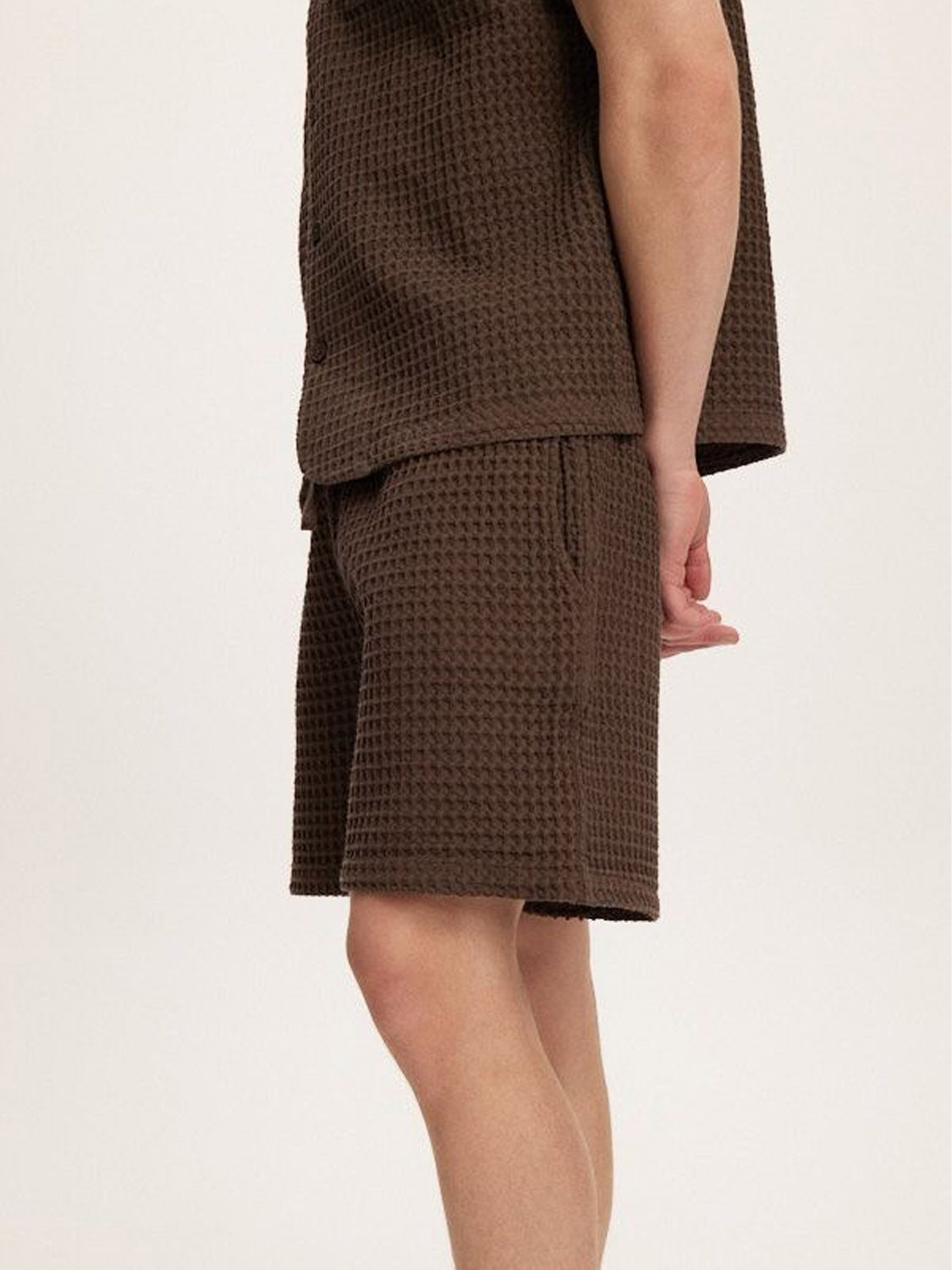 A person stands sideways against a plain light background, wearing the Kuwalla Heavy Waffle Short and matching brown relaxed fit short-sleeve shirt made of waffle-knit fabric.