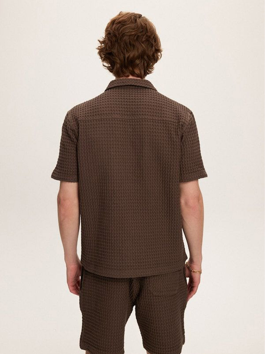 A person with wavy brown hair, seen from behind, wears Kuwalla's Heavy Waffle Short and a matching relaxed fit brown shirt made from 100% cotton. The background is plain and light-colored.