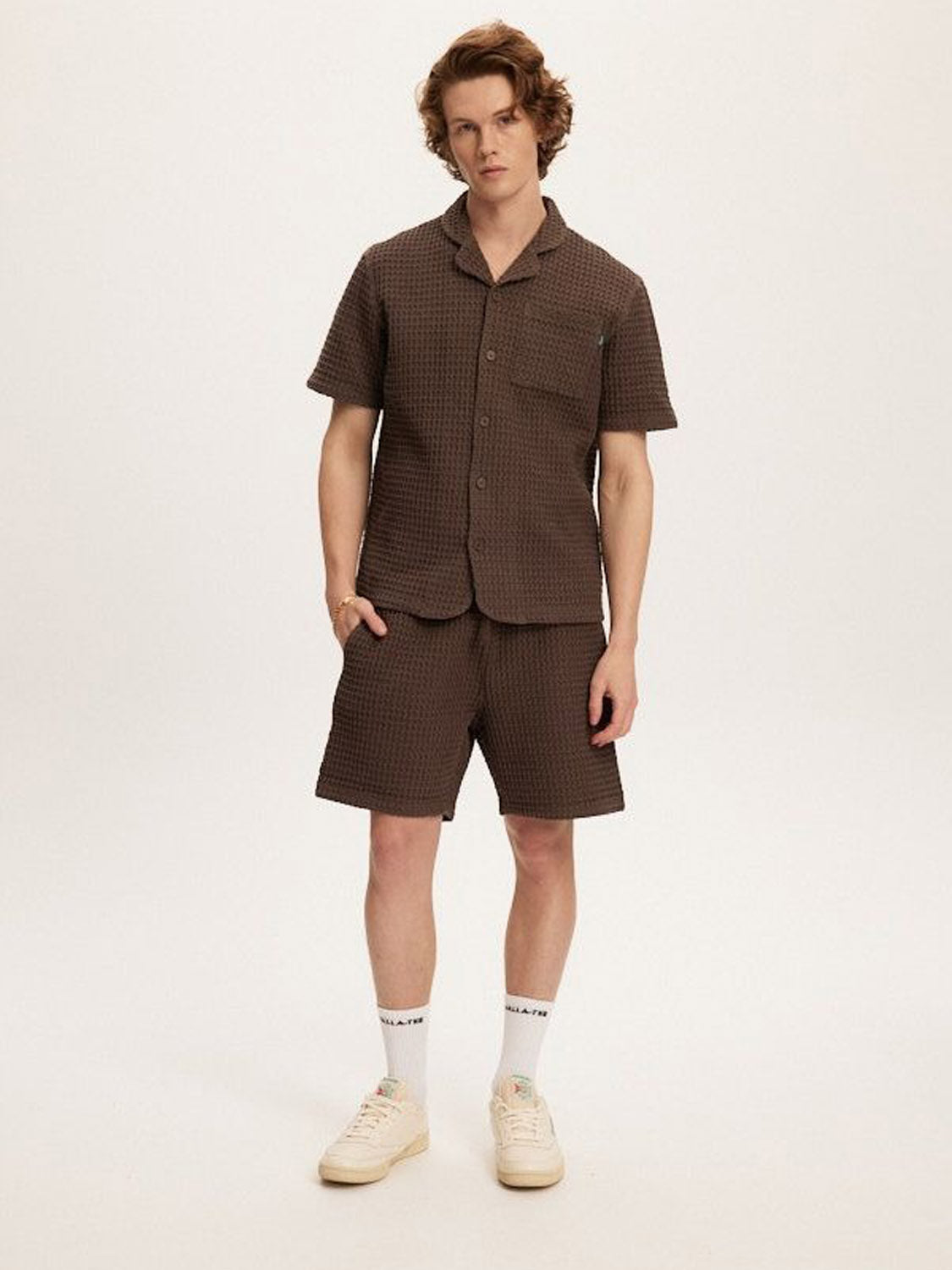 A young man with light skin and wavy brown hair wears Kuwalla's Heavy Waffle Short—a relaxed fit, brown 100% cotton short—paired with a matching button-up shirt, white socks, and sneakers against a plain background.
