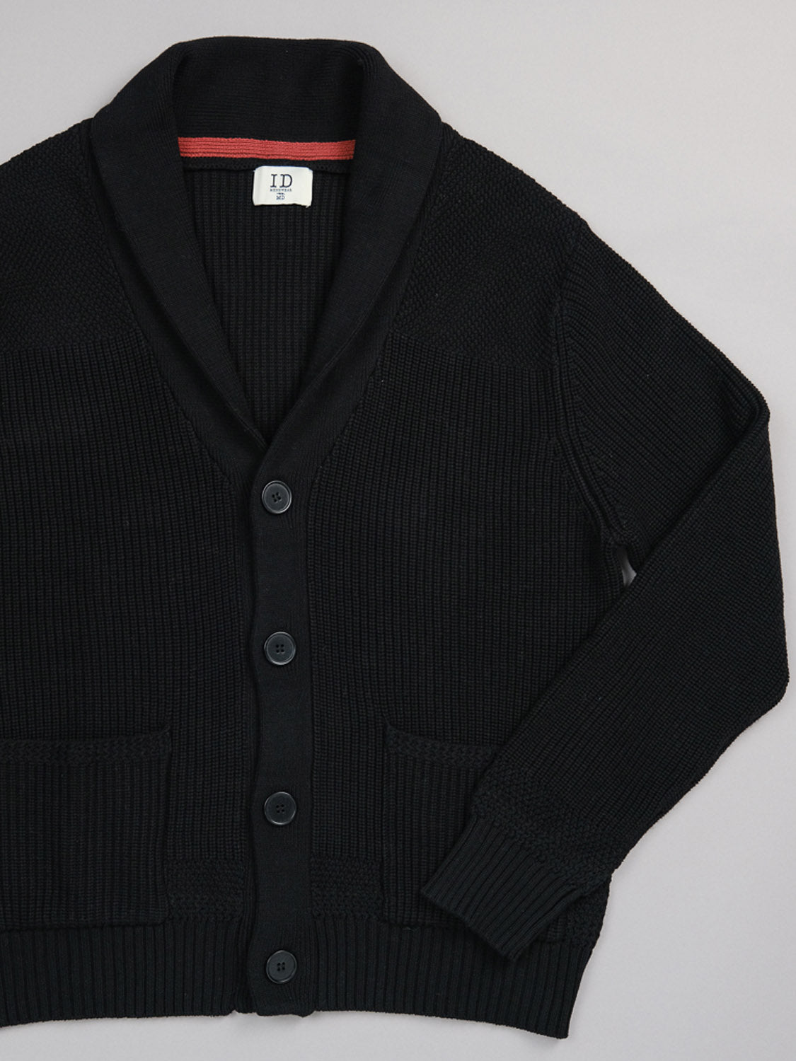 The Helsinki Cotton Knitted Cardigan from ID Menswear is made of soft cotton and includes a shawl collar, button closure, two front pockets, ribbed texture, and a small ID label at the neckline. Its shown laid flat on a plain background.