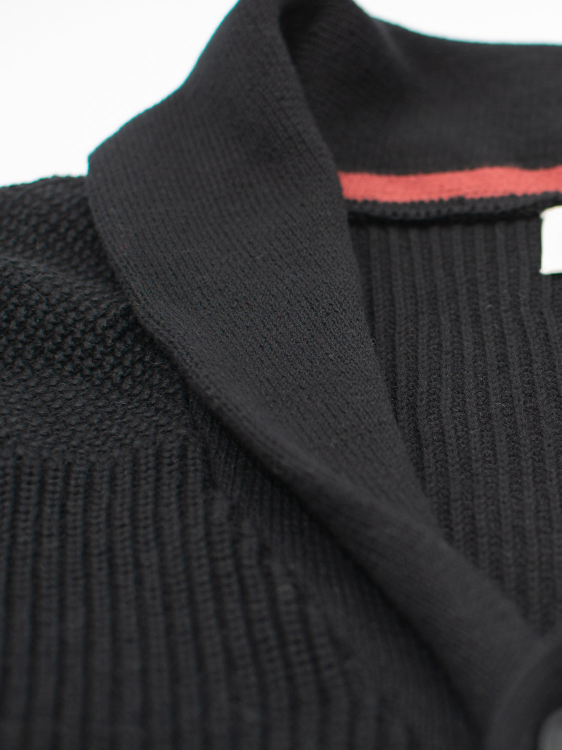 Close-up of the ID Menswear Helsinki Cotton Knitted Cardigan showcasing a classic shawl collar and black ribbed texture, with an orange stripe peeking next to the white label against a soft focus background.